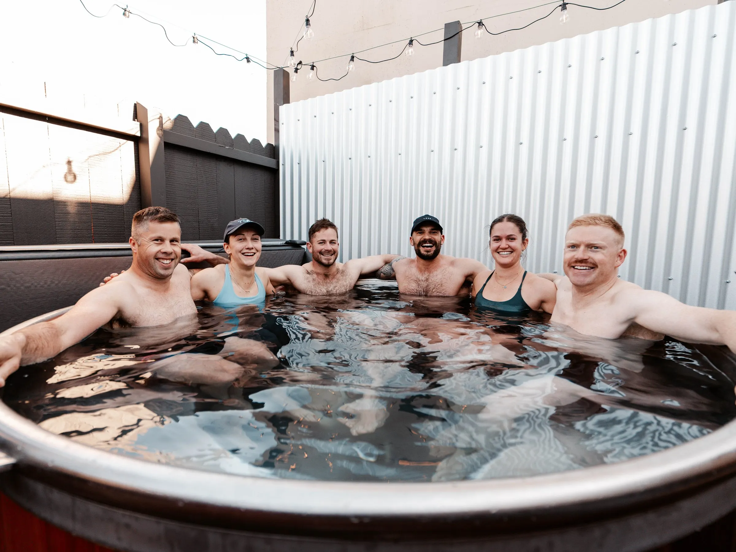 Six members shoulder to shoulder in the eight-person ice bath, all smiling at the camera.
